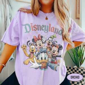 May include: A light purple t-shirt featuring the word "Disneyland" in a colorful, retro font. The shirt showcases a group of Disney characters, including Mickey Mouse, Minnie Mouse, Donald Duck, Goofy, and Pluto, in a selfie-style pose.