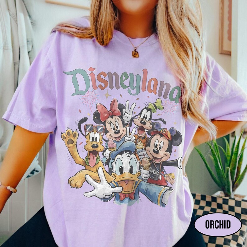 May include: A light purple t-shirt featuring the word "Disneyland" in a colorful, retro font. The shirt showcases a group of Disney characters, including Mickey Mouse, Minnie Mouse, Donald Duck, Goofy, and Pluto, in a selfie-style pose.