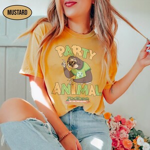 May include: Mustard-colored t-shirt featuring a cartoon sloth wearing sunglasses and a green shirt. The shirt has the words "PARTY ANIMAL" in orange and green, with "ZOOTOPIA BETTER TOGETHER" below the sloth illustration.