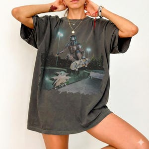 Pu&ograve; includere: Una t-shirt oversize grigio antracite con una stampa grafica di un personaggio mandaloriano in armatura che fa skateboard in una bowl, con una piccola creatura verde su uno skateboard sottostante. La maglia ha maniche corte e girocollo.