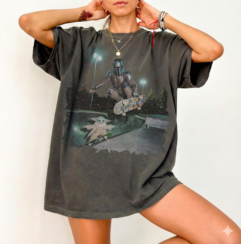 Pu&ograve; includere: Una t-shirt oversize grigio antracite con una stampa grafica di un personaggio mandaloriano in armatura che fa skateboard in una bowl, con una piccola creatura verde su uno skateboard sottostante. La maglia ha maniche corte e girocollo.