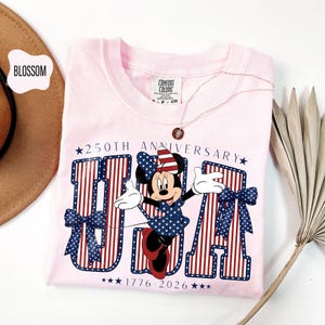 Może przedstawiać: Jasnor&oacute;żowy t-shirt z grafiką Myszki Minnie w patriotycznej kokardzie, umieszczony przed dużymi literami tworzącymi napis USA. Projekt zawiera tekst 250TH ANNIVERSARY oraz 1776-2026, z czerwono-białymi paskami i wzorami niebieskich gwiazdek.
