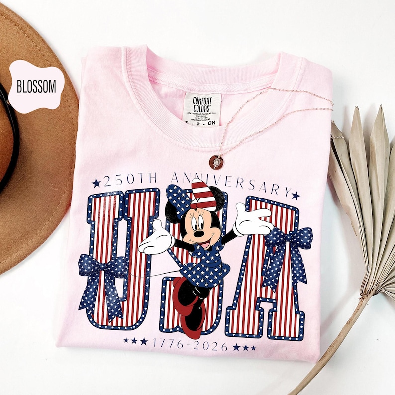 Może przedstawiać: Jasnor&oacute;żowy t-shirt z grafiką Myszki Minnie w patriotycznej kokardzie, umieszczony przed dużymi literami tworzącymi napis USA. Projekt zawiera tekst 250TH ANNIVERSARY oraz 1776-2026, z czerwono-białymi paskami i wzorami niebieskich gwiazdek.