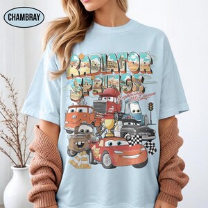May include: A light blue t-shirt featuring a graphic print of characters from the movie Cars. The design includes the text Radiator Springs and Lightning McQueen, with illustrations of various vehicles, a gold trophy, and checkered flags.