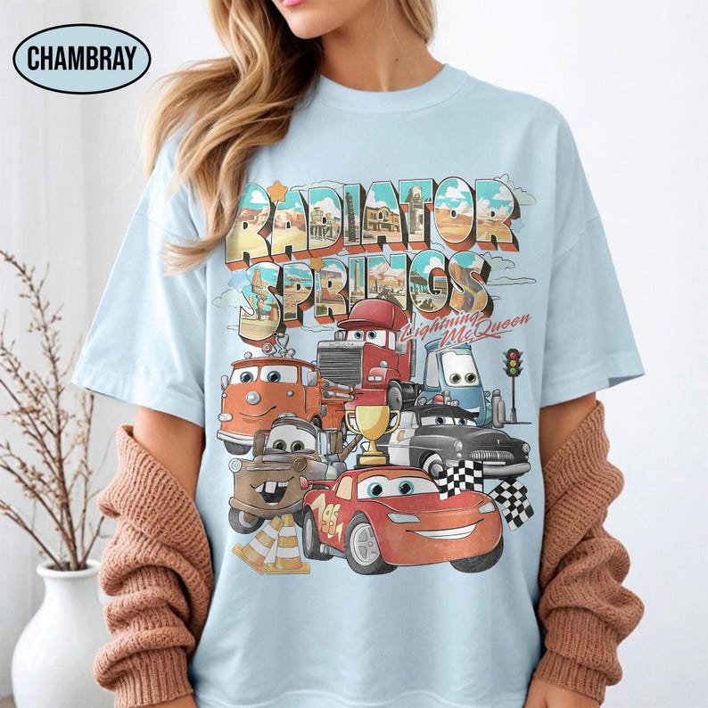 May include: A light blue t-shirt featuring a graphic print of characters from the movie Cars. The design includes the text Radiator Springs and Lightning McQueen, with illustrations of various vehicles, a gold trophy, and checkered flags.