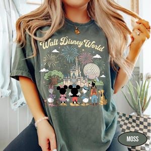 May include: Moss green t-shirt with a vibrant Walt Disney World graphic. The design showcases fireworks, a castle, Spaceship Earth, and Disney characters such as Mickey and Minnie Mouse, Donald Duck, Goofy, and Pluto. The words "Walt Disney World" are arched above.