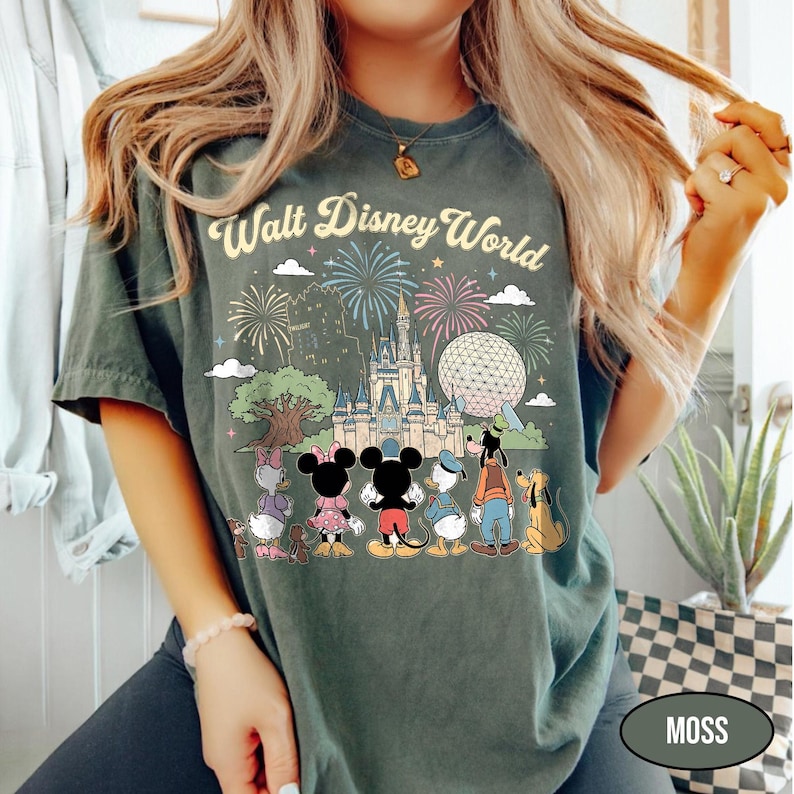May include: Moss green t-shirt with a vibrant Walt Disney World graphic. The design showcases fireworks, a castle, Spaceship Earth, and Disney characters such as Mickey and Minnie Mouse, Donald Duck, Goofy, and Pluto. The words "Walt Disney World" are arched above.