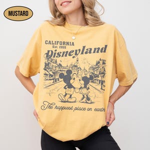May include: Mustard-colored t-shirt featuring a graphic of Mickey and Minnie Mouse walking down a street in front of Disneyland. The shirt has the words "CALIFORNIA Disneyland Est. 1955" and "The happiest place on earth."