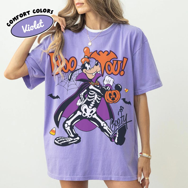 May include: A violet t-shirt featuring a graphic of Goofy dressed in a skeleton costume and a vampire cape. The text reads "BOO YOU!" in orange letters above the character, with a pumpkin, spiderweb, bats, and candy corn accents included.