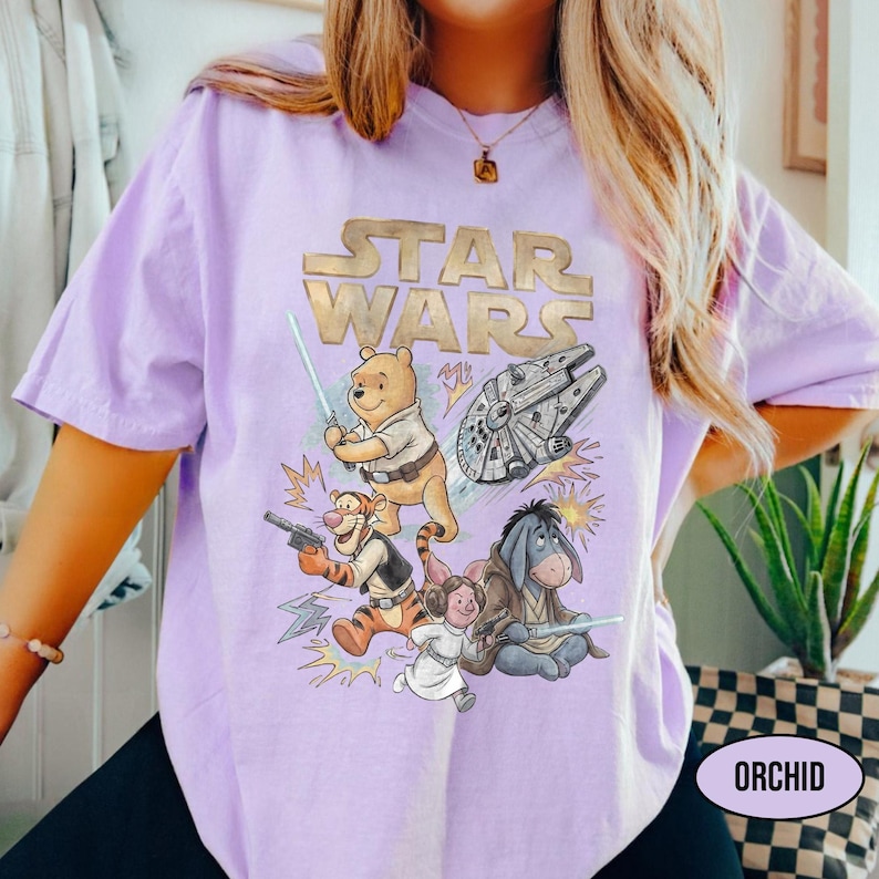 Puede incluir: Una camiseta de color lila claro con un dise&ntilde;o que combina Star Wars y los personajes de Winnie the Pooh. El dise&ntilde;o incluye a Winnie the Pooh con un sable l&aacute;ser, Tigger con un bl&aacute;ster y otros personajes. El texto "STAR WARS" est&aacute; en la parte superior.