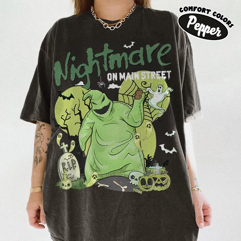 Puede incluir: Camiseta gris oscuro con un gr&aacute;fico que presenta el texto "Nightmare on Main Street" y un personaje verde de Oogie Boogie. El dise&ntilde;o incluye fantasmas, calabazas y una l&aacute;pida. La camiseta est&aacute; etiquetada como "Comfort Colors Pepper".