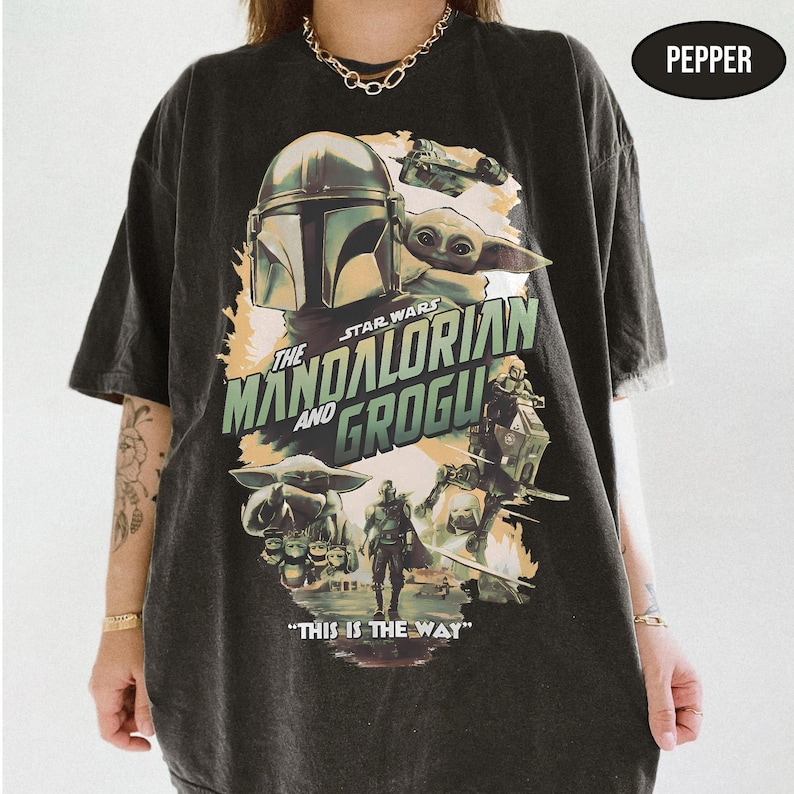 May include: Dark gray t-shirt featuring a graphic from the Star Wars series "The Mandalorian". The design includes the Mandalorian, Grogu, and the text "The Mandalorian and Grogu" and "This is the Way".