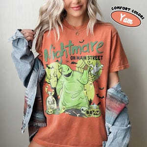 Puede incluir: Camiseta naranja con un dise&ntilde;o de "Pesadilla antes de Navidad". El dise&ntilde;o presenta al personaje Oogie Boogie, fantasmas, l&aacute;pidas y el texto "Nightmare on Main Street". La camiseta tiene un aspecto vintage.