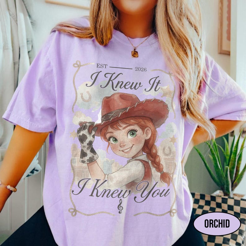 May include: A light purple t-shirt featuring a graphic of a cartoon character wearing a brown cowboy hat and a cow-print glove. The text reads EST 2026, I Knew It, and I Knew You, framed by a decorative rope border with stars and horseshoes.