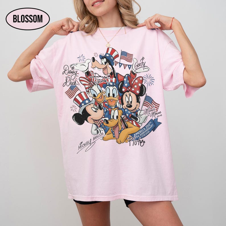 Może przedstawiać: Jasnor&oacute;żowa koszulka z grafiką postaci Disneya, w tym Myszki Miki i Myszki Minnie, w patriotycznych strojach. Projekt zawiera amerykańskie flagi i fajerwerki, z tekstem "America 250th Anniversary 1776-2026".