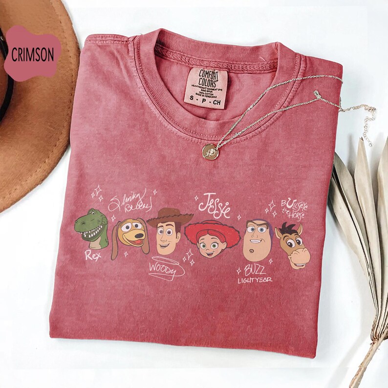 May include: A crimson short-sleeved t-shirt featuring a graphic print of Toy Story character heads including Rex, Slinky Dog, Woody, Jessie, Buzz Lightyear, and Bullseye. The shirt has a crew neckline and a Comfort Colors brand label at the collar.