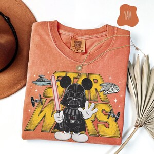 Puede incluir: Camiseta naranja Comfort Colors con un gr&aacute;fico de Darth Vader con orejas de Mickey Mouse, sosteniendo un sable de luz y el logo de Star Wars. La camiseta incluye im&aacute;genes del Halc&oacute;n Milenario y cazas TIE. Tambi&eacute;n se ven un collar dorado y un sombrero marr&oacute;n.