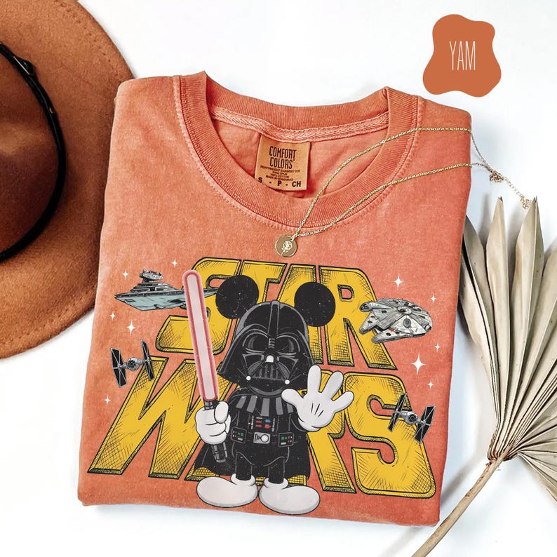 Puede incluir: Camiseta naranja Comfort Colors con un gr&aacute;fico de Darth Vader con orejas de Mickey Mouse, sosteniendo un sable de luz y el logo de Star Wars. La camiseta incluye im&aacute;genes del Halc&oacute;n Milenario y cazas TIE. Tambi&eacute;n se ven un collar dorado y un sombrero marr&oacute;n.