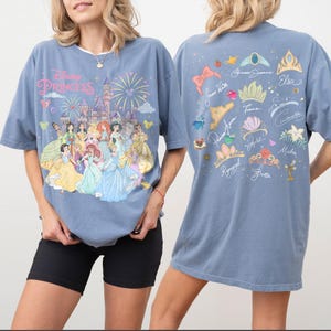 K&ouml;nnte beinhalten: Ein blaues T-Shirt mit kurzen &Auml;rmeln, das auf der Vorderseite Disney-Prinzessinnen vor einem Schloss mit Feuerwerk und den Schriftzug Disney Princess zeigt. Die R&uuml;ckseite zeigt verschiedene Kronen und Accessoires mit den Namen der Figuren.