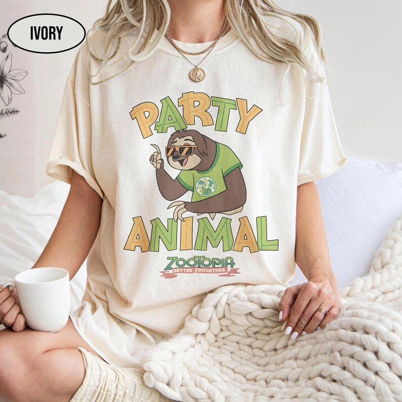 May include: Ivory-colored t-shirt featuring a cartoon sloth wearing sunglasses and a green shirt. The shirt has the words "PARTY ANIMAL" in orange and green, with "ZOOTOPIA BETTER ZOOGETHER" below the sloth.