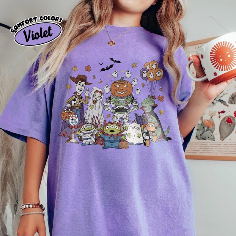 May include: A violet Comfort Colors t-shirt with a Halloween-themed cartoon graphic. The design features a pumpkin, ghosts, bats, and other characters. The shirt has a relaxed fit and short sleeves, perfect for autumn.