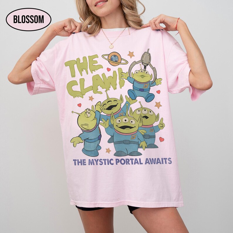 May include: Light pink t-shirt featuring a graphic of green aliens with the text "THE CLAW!" and "THE MYSTIC PORTAL AWAITS". The design includes a planet, stars, and a claw machine. The shirt has a relaxed fit.