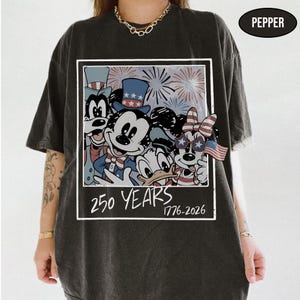 May include: A charcoal gray t-shirt featuring a graphic print of cartoon characters wearing patriotic hats and star-shaped glasses. The design includes fireworks, a small American flag, and the text 250 YEARS 1776-2026 in a white rectangular frame.