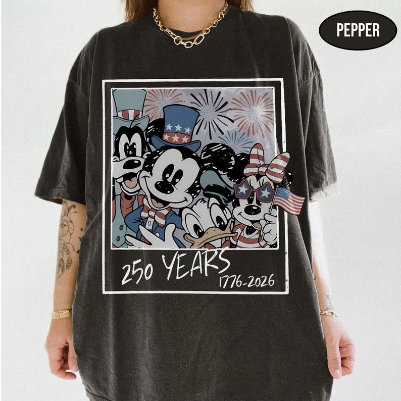 May include: A charcoal gray t-shirt featuring a graphic print of cartoon characters wearing patriotic hats and star-shaped glasses. The design includes fireworks, a small American flag, and the text 250 YEARS 1776-2026 in a white rectangular frame.