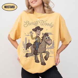 May include: A mustard yellow short-sleeve t-shirt featuring a graphic print of Sheriff Woody riding a horse. The text "Sheriff Woody" is printed above the character. The design includes a desert landscape with cacti and skulls in the background.