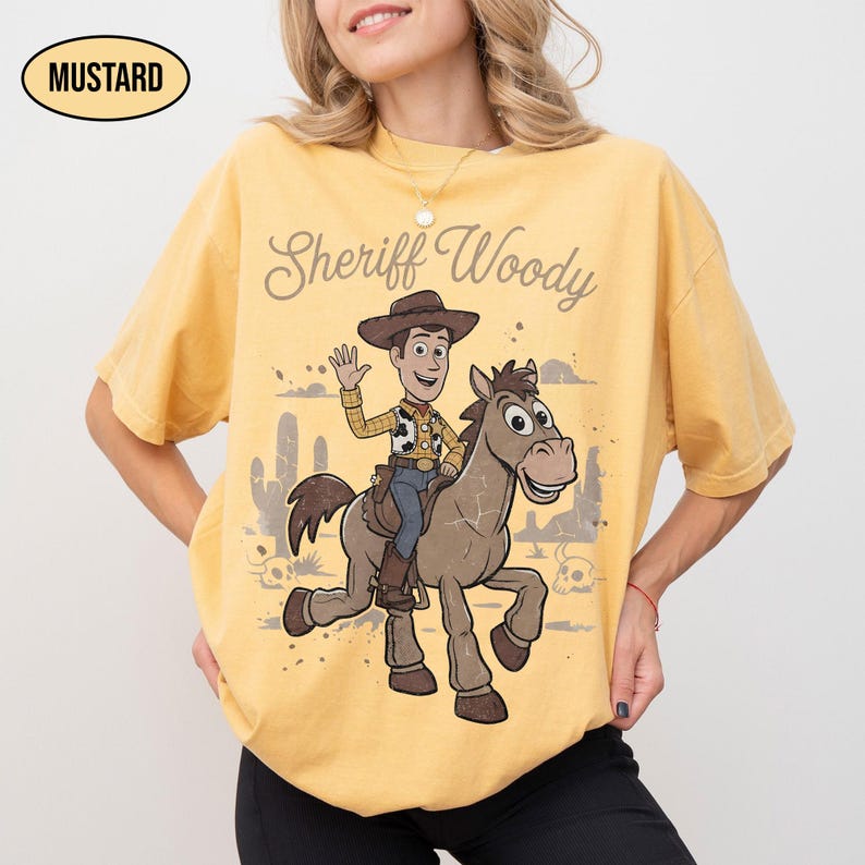 May include: A mustard yellow short-sleeve t-shirt featuring a graphic print of Sheriff Woody riding a horse. The text "Sheriff Woody" is printed above the character. The design includes a desert landscape with cacti and skulls in the background.