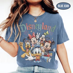 May include: Blue jean colored t-shirt featuring the word "Disneyland" in colorful lettering. The shirt displays a graphic of Disney characters including Mickey Mouse, Minnie Mouse, Donald Duck, Goofy, and Pluto. The shirt has a crew neck.
