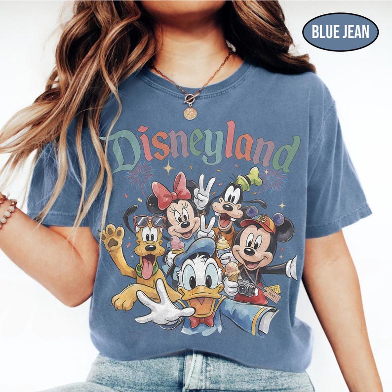 May include: Blue jean colored t-shirt featuring the word "Disneyland" in colorful lettering. The shirt displays a graphic of Disney characters including Mickey Mouse, Minnie Mouse, Donald Duck, Goofy, and Pluto. The shirt has a crew neck.