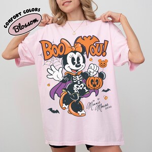 May include: A light pink t-shirt featuring a graphic of Minnie Mouse dressed in a skeleton costume with a purple cape, holding a pumpkin bucket. The text reads Boo You! and Minnie Mouse xoxo, with spiderwebs, bats, and candy corn accents.
