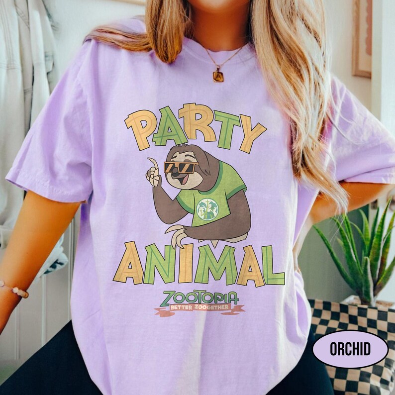 May include: Orchid-colored t-shirt featuring a cartoon sloth wearing sunglasses and a green shirt. The shirt has the words "PARTY ANIMAL" in yellow and green, with "ZOOTOPIA BETTER ZOO TOGETHER" below the sloth graphic.