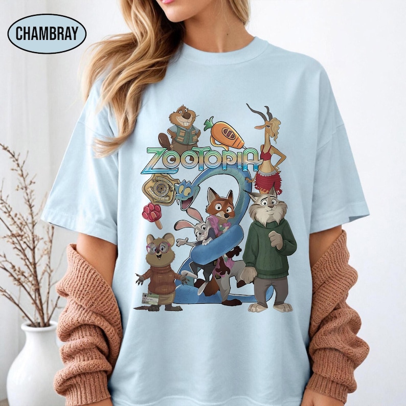 May include: Light blue t-shirt featuring the word "Zootopia" and cartoon characters from the movie. The graphic includes a fox, rabbit, and other animals. The shirt is made of a soft material.