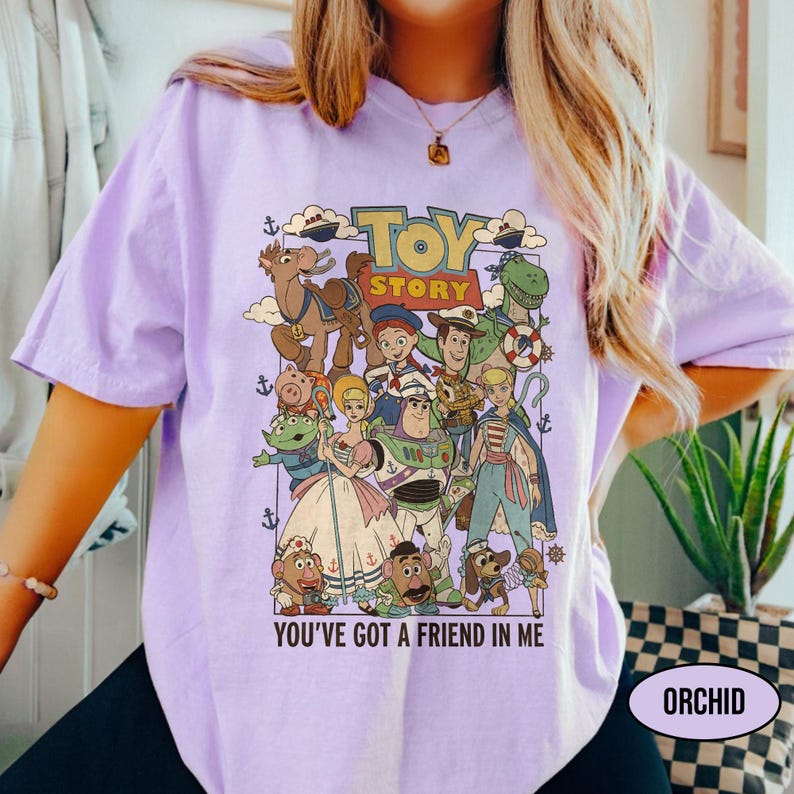 May include: A light purple t-shirt featuring a colorful graphic of characters from the animated film "Toy Story." The design includes the text "TOY STORY" and "YOU'VE GOT A FRIEND IN ME." The shirt also has the word "ORCHID" printed on it.