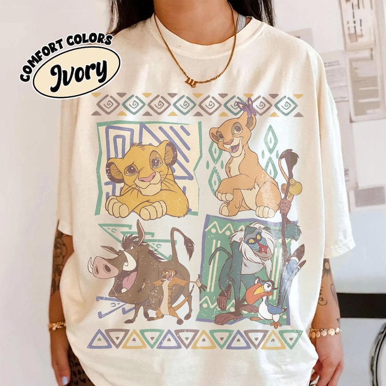 May include: An ivory t-shirt featuring a graphic print of characters from The Lion King, including Simba, Nala, Pumbaa, Timon, Rafiki, and Zazu, arranged in four rectangular panels with geometric borders and the text COMFORT COLORS IVORY.