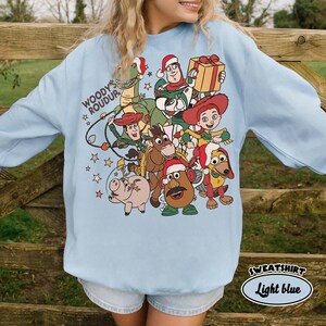 Op de afbeelding: Lichtblauwe sweatshirt met een feestelijke afbeelding van Toy Story-personages met kerstmutsen en cadeaus. De tekst "WOODY & ROUDYR" staat boven de personages. De tekst "SWEATSHIRT Light blue" staat rechtsonder.