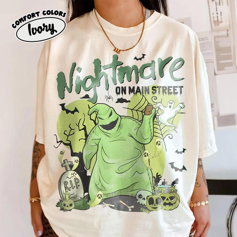 Puede incluir: Camiseta color marfil con un gr&aacute;fico verde que dice "Nightmare on Main Street". El dise&ntilde;o presenta un personaje verde, fantasmas, murci&eacute;lagos y calabazas. La camiseta tiene un corte holgado y mangas cortas.