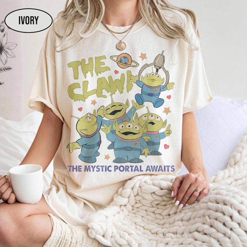 May include: Ivory-colored t-shirt featuring a cartoon graphic of green aliens in blue suits, with the text "THE CLAW!" and "THE MYSTIC PORTAL AWAITS." The design includes stars, a planet, and a claw machine. The shirt is short-sleeved.