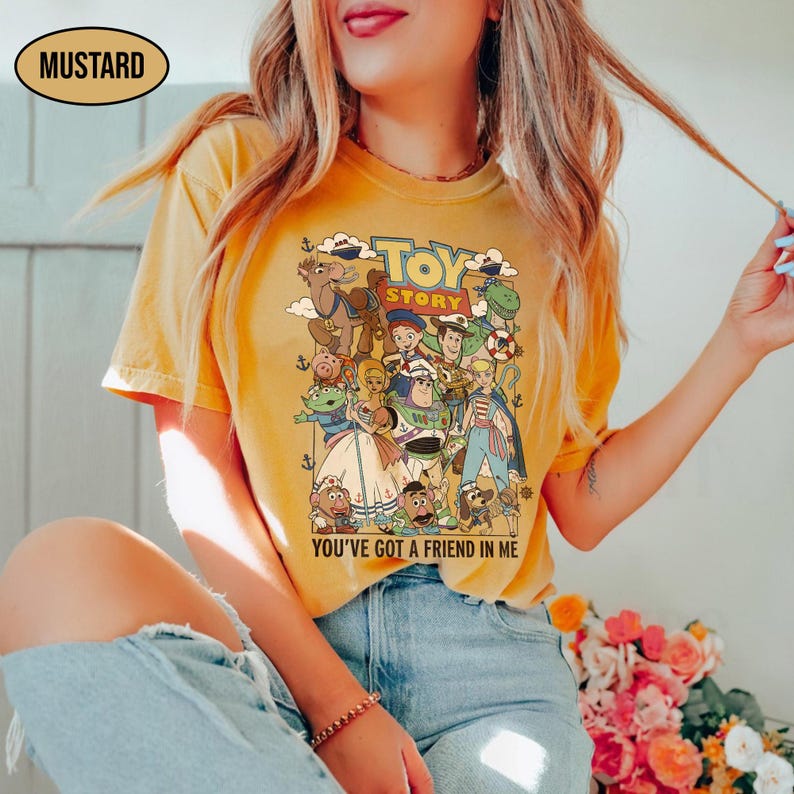 May include: Mustard-colored t-shirt featuring a graphic of characters from the animated film "Toy Story." The design includes the text "TOY STORY" and "YOU'VE GOT A FRIEND IN ME." The shirt is a casual style.