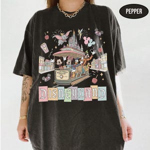 May include: Black t-shirt featuring a Disneyland design. The graphic includes a Main Street Trolley with Mickey, Minnie, Donald, and Goofy, along with Dumbo, fireworks, and the Disneyland logo in pastel colors. The shirt has a relaxed fit.