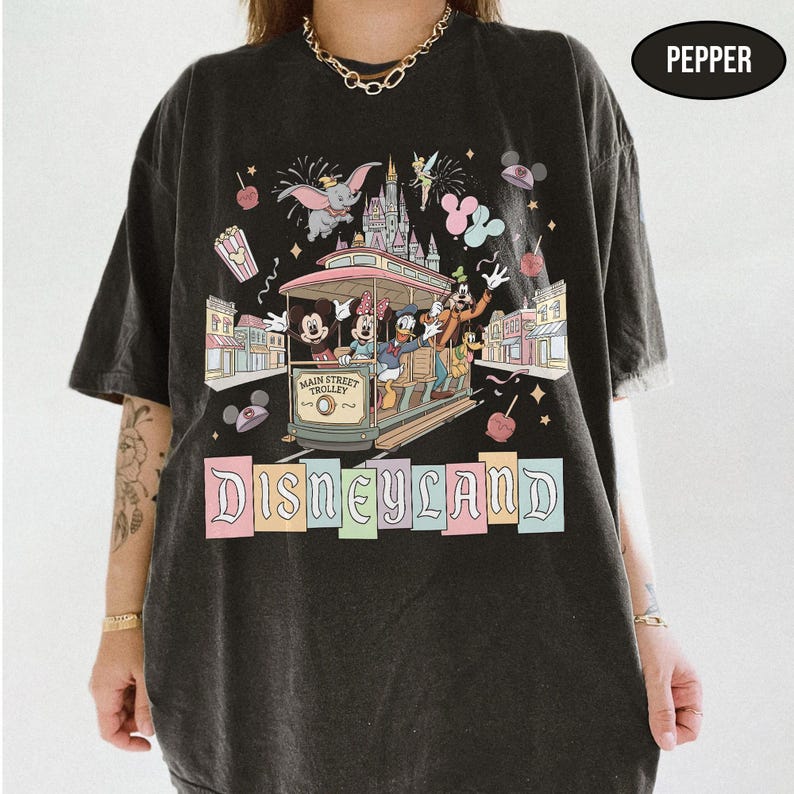May include: Black t-shirt featuring a Disneyland design. The graphic includes a Main Street Trolley with Mickey, Minnie, Donald, and Goofy, along with Dumbo, fireworks, and the Disneyland logo in pastel colors. The shirt has a relaxed fit.