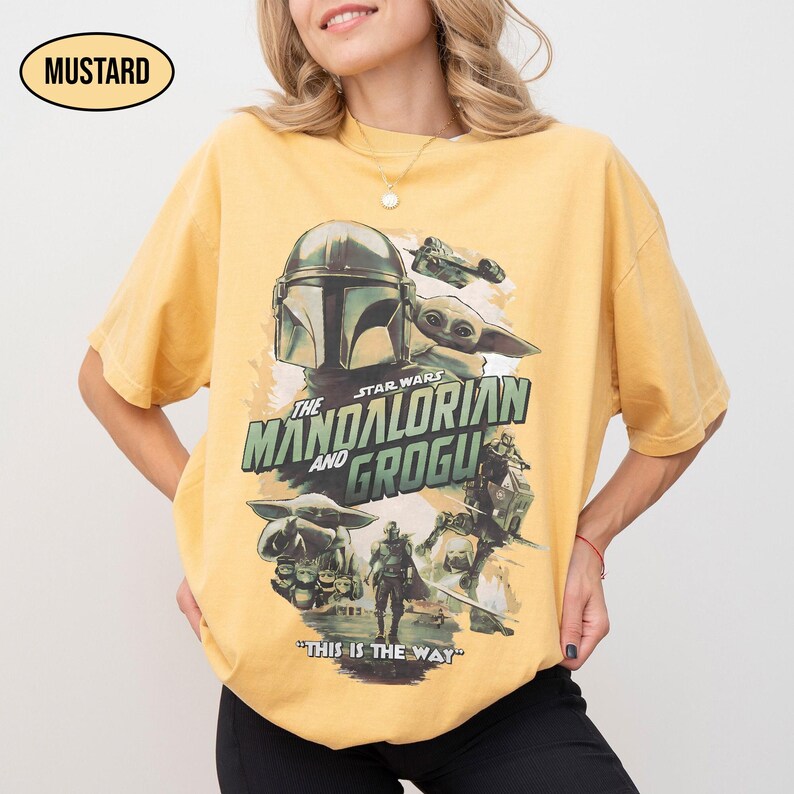 May include: Mustard-colored t-shirt featuring a graphic from the Star Wars series, "The Mandalorian and Grogu." The design includes the Mandalorian helmet, Grogu, and the phrase "This is the Way."