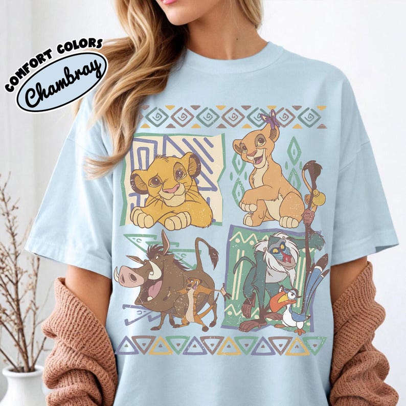 May include: A light blue t-shirt featuring a graphic print of characters from The Lion King, including Simba, Nala, Pumbaa, Timon, Rafiki, and Zazu. The design includes geometric patterns and the text Comfort Colors Chambray in the upper corner.