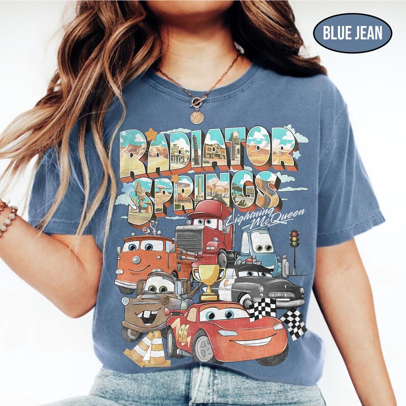 May include: A blue jean colored t-shirt featuring a graphic print of Radiator Springs with Lightning McQueen and other characters from the movie. The design includes the text Radiator Springs and Lightning McQueen with a gold trophy.