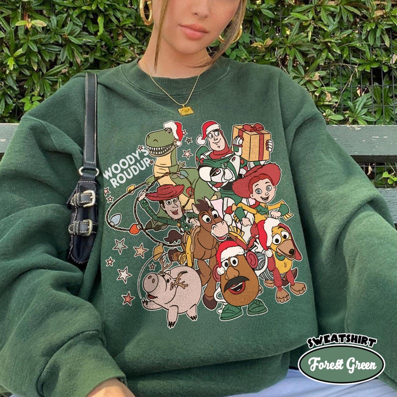 Op de afbeelding: Bostgroen sweatshirt met een afbeelding van Toy Story-personages met kerstmutsen. Het ontwerp bevat Woody, Buzz Lightyear en andere personages. De tekst "WOODY'S ROUDUP" staat links en "SWEATSHIRT Forest Green" rechtsonder.