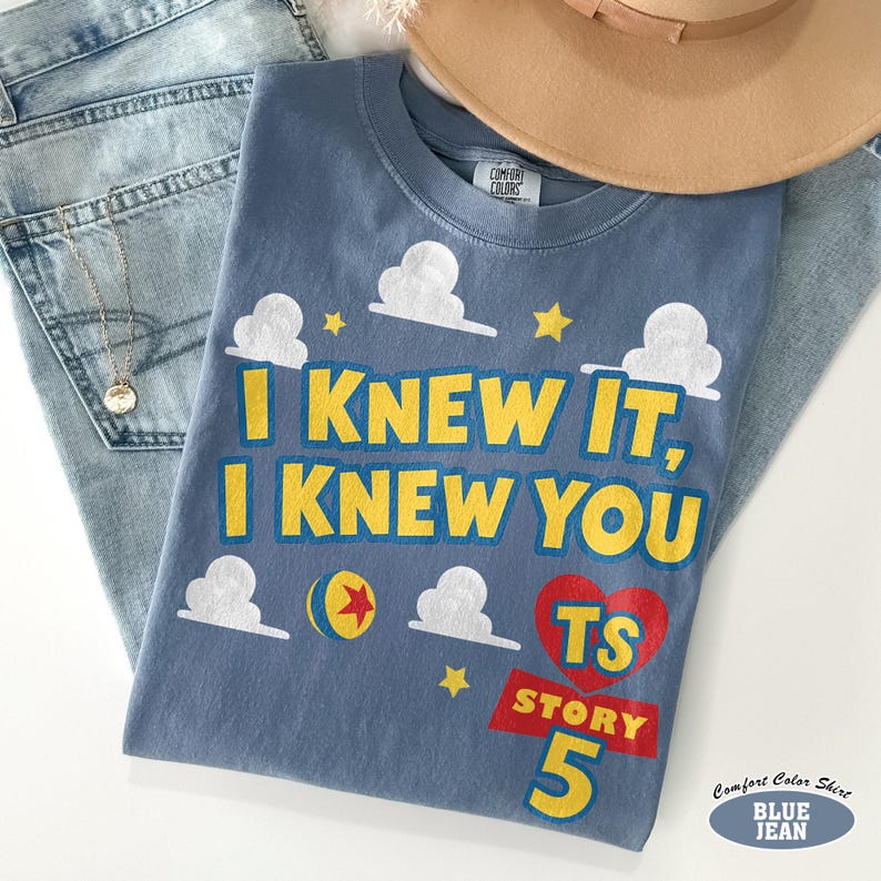 Puede incluir: Una camiseta azul con el texto amarillo I KNEW IT, I KNEW YOU, gr&aacute;ficos de nubes blancas, estrellas amarillas, una pelota colorida y un coraz&oacute;n rojo que contiene el texto amarillo TS STORY 5. La camiseta tiene una etiqueta Comfort Colors.