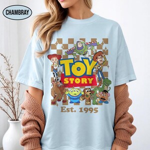 May include: Light blue t-shirt featuring a colorful graphic of Toy Story characters, including Woody, Buzz Lightyear, and others. The words "TOY STORY" are in yellow and red, with "Est. 1995" below. The shirt has a crew neck.