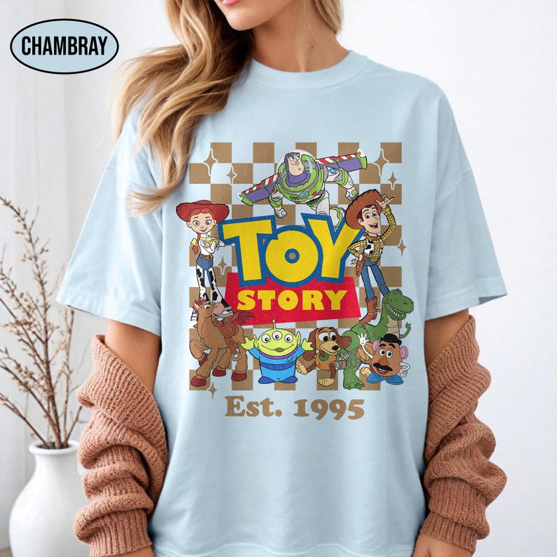 May include: Light blue t-shirt featuring a colorful graphic of Toy Story characters, including Woody, Buzz Lightyear, and others. The words "TOY STORY" are in yellow and red, with "Est. 1995" below. The shirt has a crew neck.